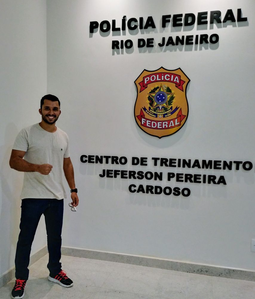 Filipe sorrindo, de punho fechado, ao lado do brasão da Polícia Federal no Centro de Treinamento do Rio de Janeiro